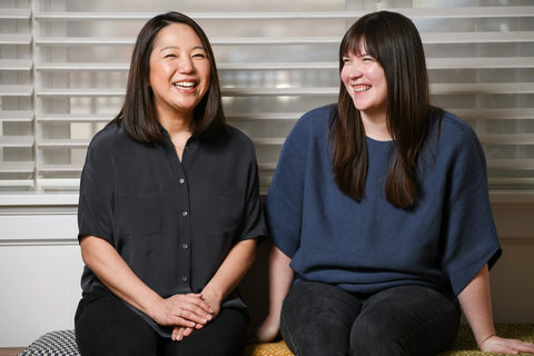 Two women sitting together smiling in a room with striped wallpaper.
