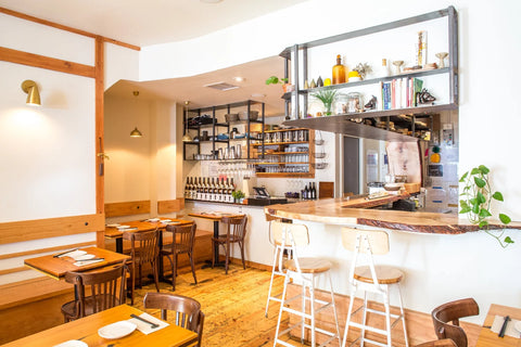 Modern restaurant interior with wooden tables, chairs, and shelves stocked with bottles.